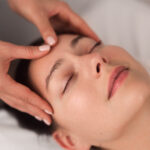 From above of crop unrecognizable cosmetician massaging forehead of young female client lying on couch with closed eyes during facial procedure in light salon